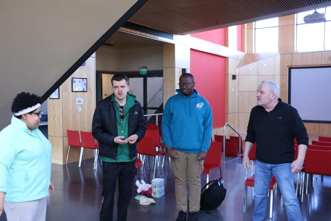 Four CreatAbility participants standing in a loose circle mid-scene in a bright community room, listening to the person who is speaking.